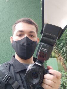 Fotógrafo do IDV News usando máscara e segurando câmera profissional com flash externo em área externa arborizada.