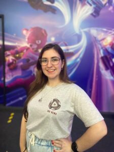 Colaboradora do IDV News sorrindo em frente a painel colorido de evento de games, vestindo camiseta personalizada.