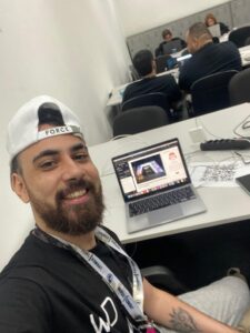 Colaborador do IDV News sorrindo enquanto trabalha em laptop na área de imprensa de evento, vestindo boné branco.