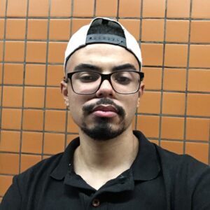 Homem de boné e óculos posa para foto em frente a parede de azulejos laranja, usando camiseta polo preta.