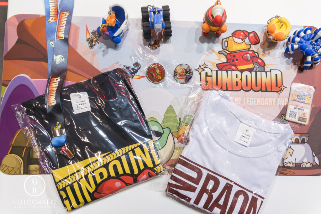 Gravity na Gamescom Latam exibe brindes de Gunbound como camisetas, chaveiros, bottons e miniaturas sobre mousepad temático.