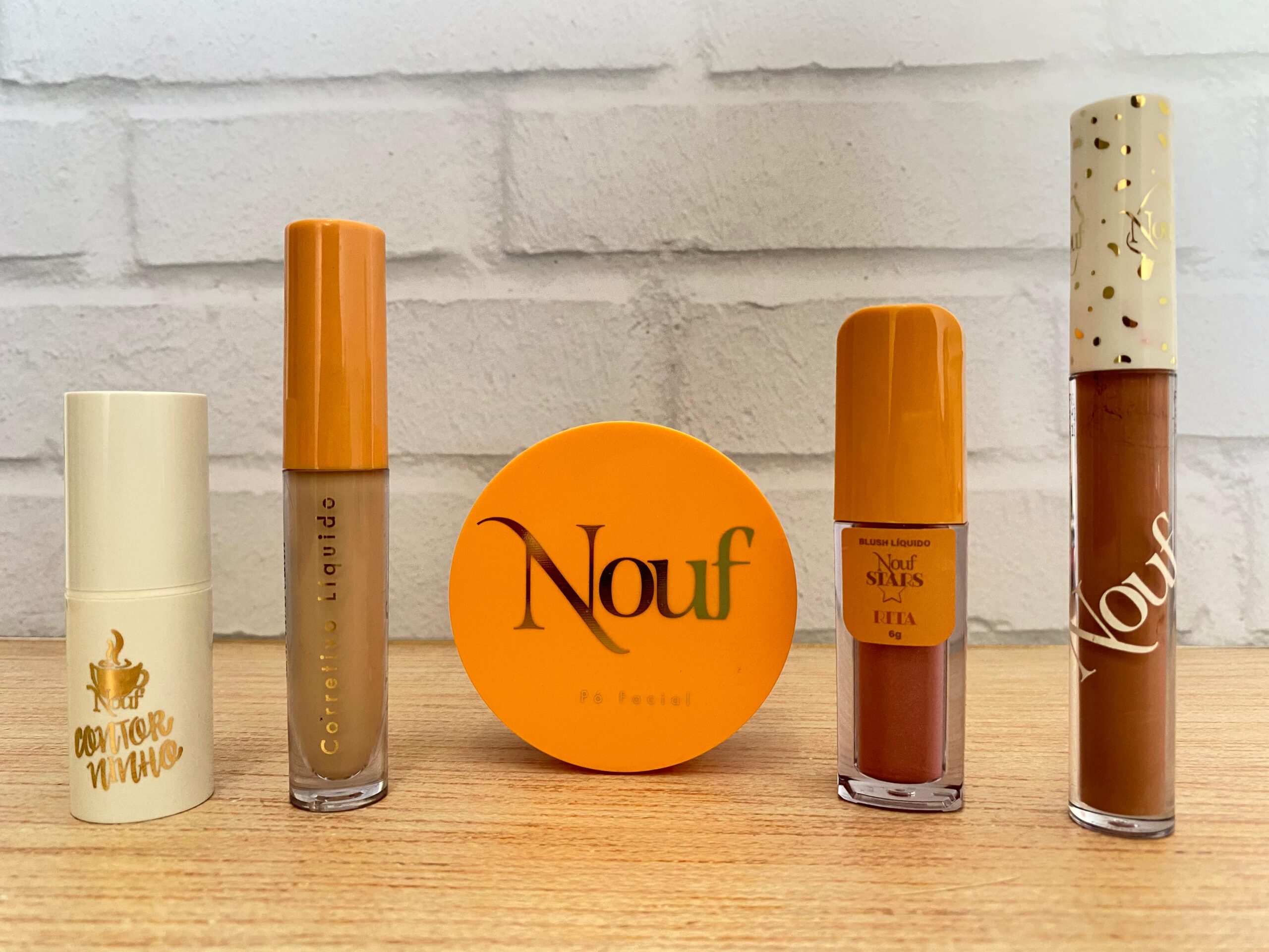 Nouf apresenta linha completa de maquiagem com cinco produtos dispostos lado a lado: bastão contorno “contorninho” em embalagem bege, corretivo líquido com tampa laranja, pó facial compacto com estojo redondo laranja, blush líquido Nouf Stars na cor Rita e gloss labial nude com tampa branca decorada.