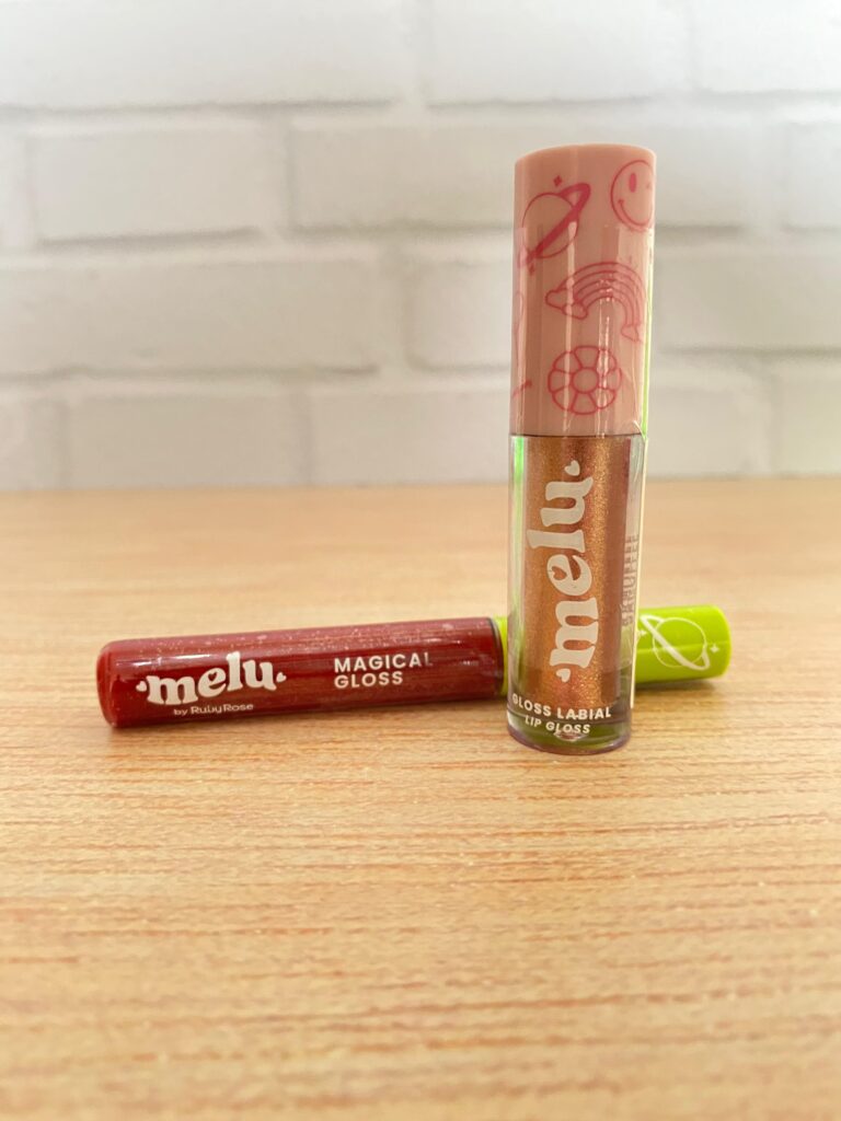 Gloss labial Ruby Rose da linha Melu com acabamento cintilante e embalagem colorida. A versão "Magical Gloss" vem em dois tons vibrantes com tampas decoradas.