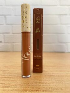 Nouf gloss labial da linha Gelateria na cor Doce de Leite, com embalagem transparente, tampa branca com detalhes dourados e caixinha marrom decorada, contendo 4 ml.