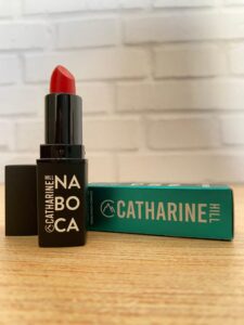 Catharine Hill batom vermelho semimate da linha Na Boca, em embalagem preta com escrita branca e caixa azul turquesa ao fundo, com logo da marca em destaque.