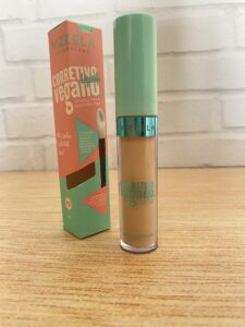Vizzela corretivo vegano em frasco transparente com tampa verde-água e texto azul. A caixa laranja e verde exibe o aplicador e informa “alta cobertura” com acabamento matte.