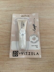 Vizzela balm labial Sugar Balm em embalagem plástica no tom creme, com estampa de cupcake e temática vanilla cake. Produto apresentado em cartela bege decorada com ilustrações e a marca Zaxy + Vizzela.