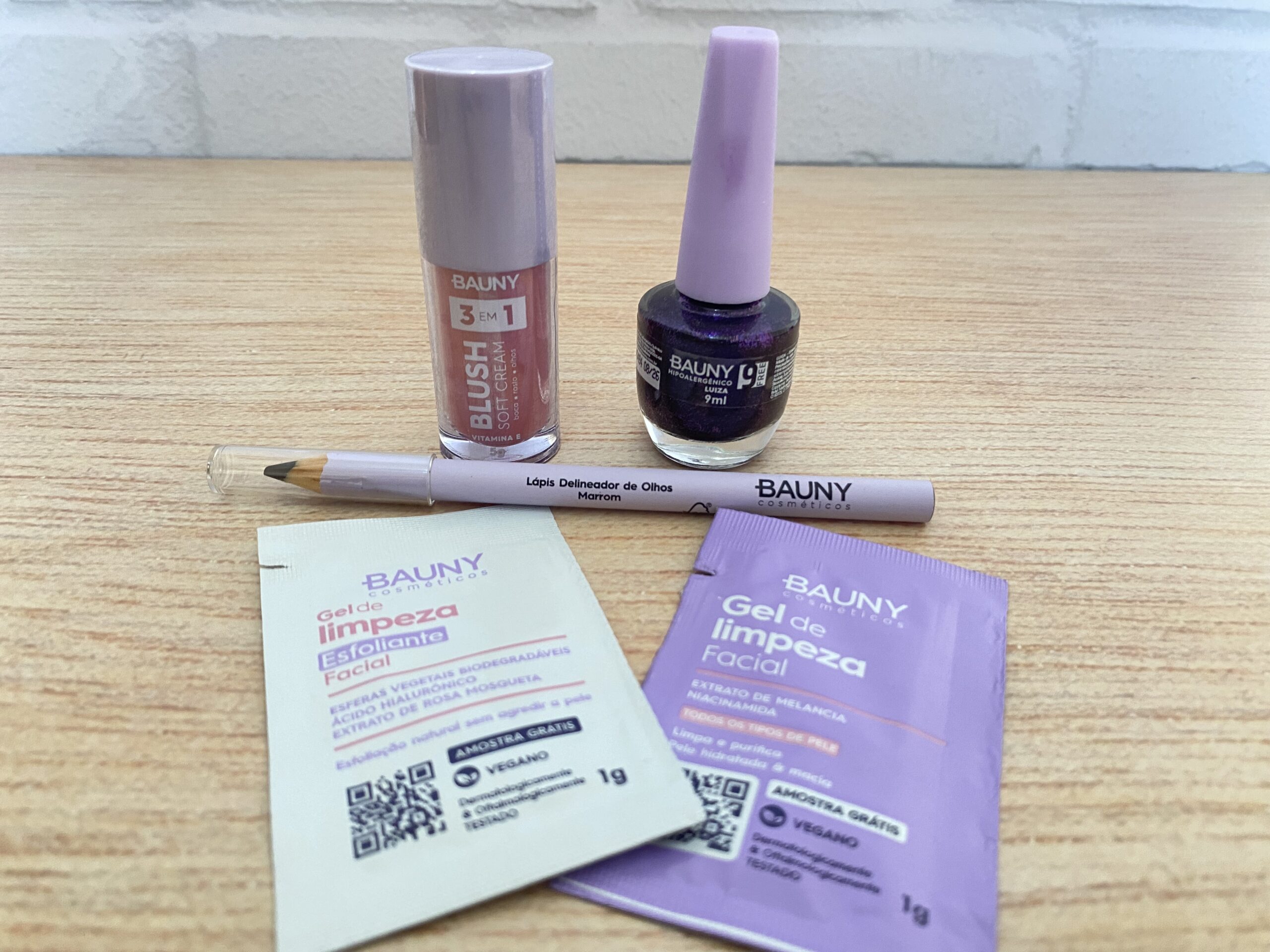 Bauny exibe cinco produtos sobre uma superfície de madeira clara: blush 3 em 1 em embalagem cilíndrica rosa, esmalte roxo cintilante com tampa lilás, lápis delineador marrom com tampa transparente e dois sachês de gel de limpeza facial, um esfoliante bege e outro hidratante lilás.