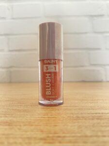 Bauny blush cremoso 3 em 1 com vitamina E, em embalagem cilíndrica transparente com tampa rosa metálica. Produto multifuncional indicado para lábios, bochechas e olhos.