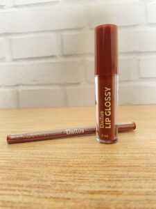 Dailus Lip Glossy na cor marrom, em frasco transparente de 3 ml com tampa laranja. O produto aparece ao lado de um lápis labial marrom com o nome Dailus em branco.