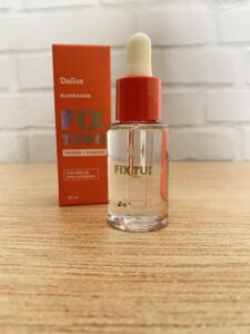Dailus Fix Tudo primer e fixador em frasco transparente com conta-gotas e tampa laranja. Ao fundo, a caixinha laranja destaca a função “blindagem” e a presença de óleo de rosa mosqueta.