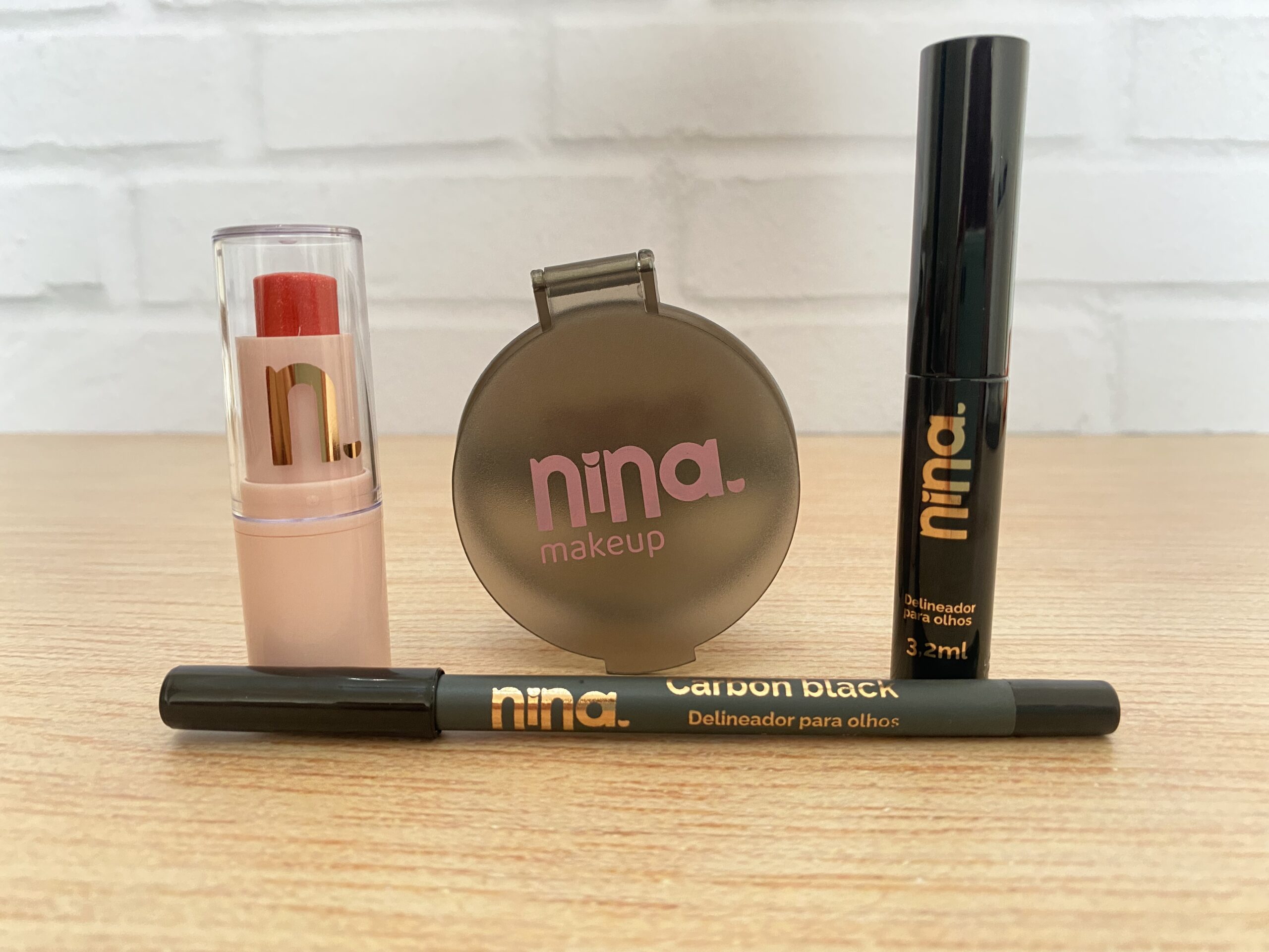 Nina MakeUp apresenta quatro itens de maquiagem sobre superfície de madeira clara: um balm labial vermelho em bastão com embalagem nude e tampa transparente, um espelho redondo marrom com logo rosa, um delineador líquido preto com frasco cilíndrico e um lápis delineador Carbon Black com tampa preta e corpo grafite. Os produtos têm visual moderno e identidade jovem.