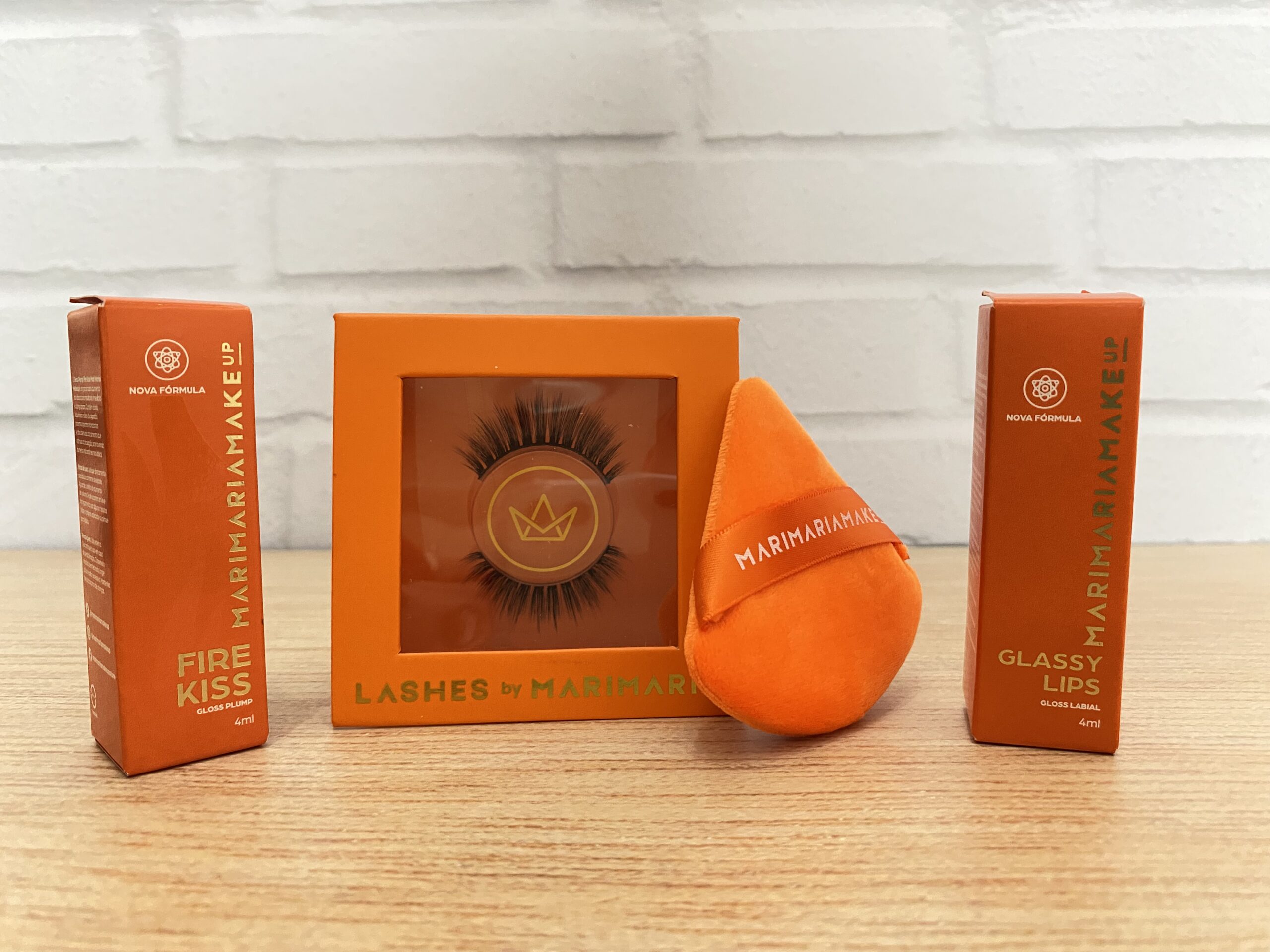 Mari Maria MakeUp apresenta kit com embalagem laranja vibrante contendo dois glosses labiais — Fire Kiss e Glassy Lips — ambos com nova fórmula em caixinhas verticais de 4 ml. Ao centro, cílios postiços volumosos na embalagem “Lashes by Mari Maria” e, ao lado, esponja de maquiagem aveludada em formato gota com fita personalizada da marca. Todos os itens estão dispostos sobre uma superfície de madeira clara, reforçando a identidade visual marcante da Mari Maria MakeUp.