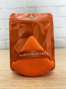 Mari Maria MakeUp esponja de maquiagem em formato de gota, cor laranja vibrante, com faixa personalizada e embalagem plástica transparente do mesmo tom.