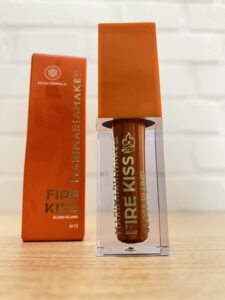 Mari Maria MakeUp gloss labial Fire Kiss, com embalagem transparente, tampa laranja e detalhes dourados. Ao fundo, caixinha laranja com o nome em dourado.