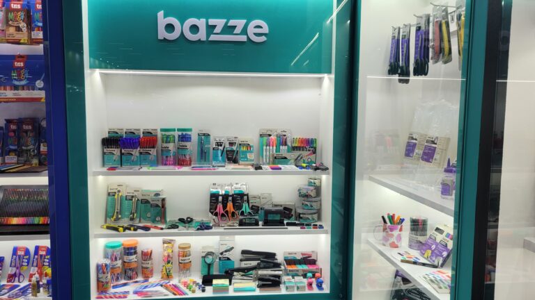 Tris compartilha espaço com a marca Bazze, que exibe itens de escritório e papelaria como canetas, marcadores, grampeadores, tesouras e organizadores, com foco em funcionalidade e design moderno.