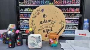 Posca apresenta painel com a frase “O amor mora aqui” pintada em MDF, ao lado de canecas, marcadores coloridos e objetos personalizados, demonstrando a aplicação criativa dos marcadores em superfícies variadas.
