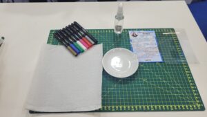 Tris compartilha espaço com a marca Bazze em uma mesa preparada para pintura em porcelana, com marcadores especiais, instruções passo a passo e utensílios para personalização de pratos e cerâmicas.