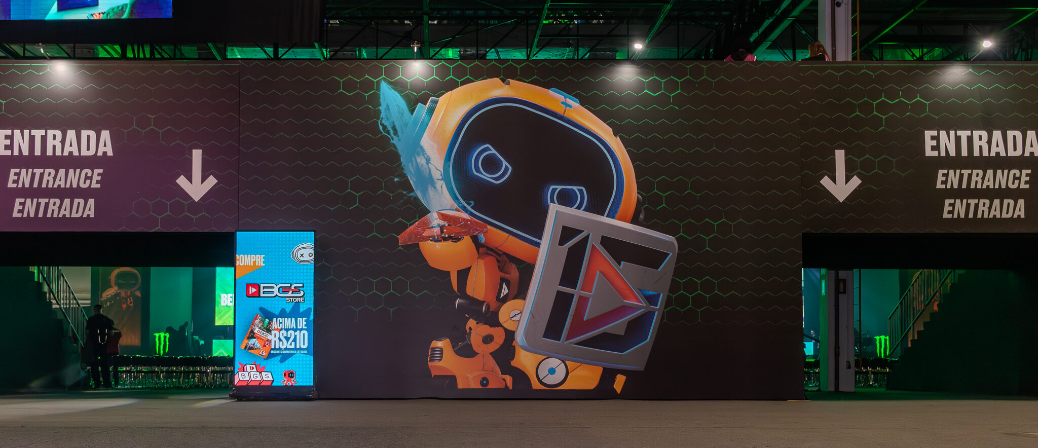 BGS: Entrada principal da Brasil Game Show com arte futurista do mascote oficial. Estrutura imersiva com iluminação verde, telão gamer e sinalização bilíngue destacam o clima do maior evento de games da América Latina.