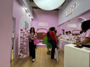 Estande da Nina Makeup na Beauty Fair 2025 em tons de rosa, com consumidores explorando lançamentos e atendentes apresentando os produtos da marca.