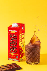 Achocolatado vegetal Possible com menos açúcar, exibido ao lado de copo com splash de bebida e barra de chocolate.