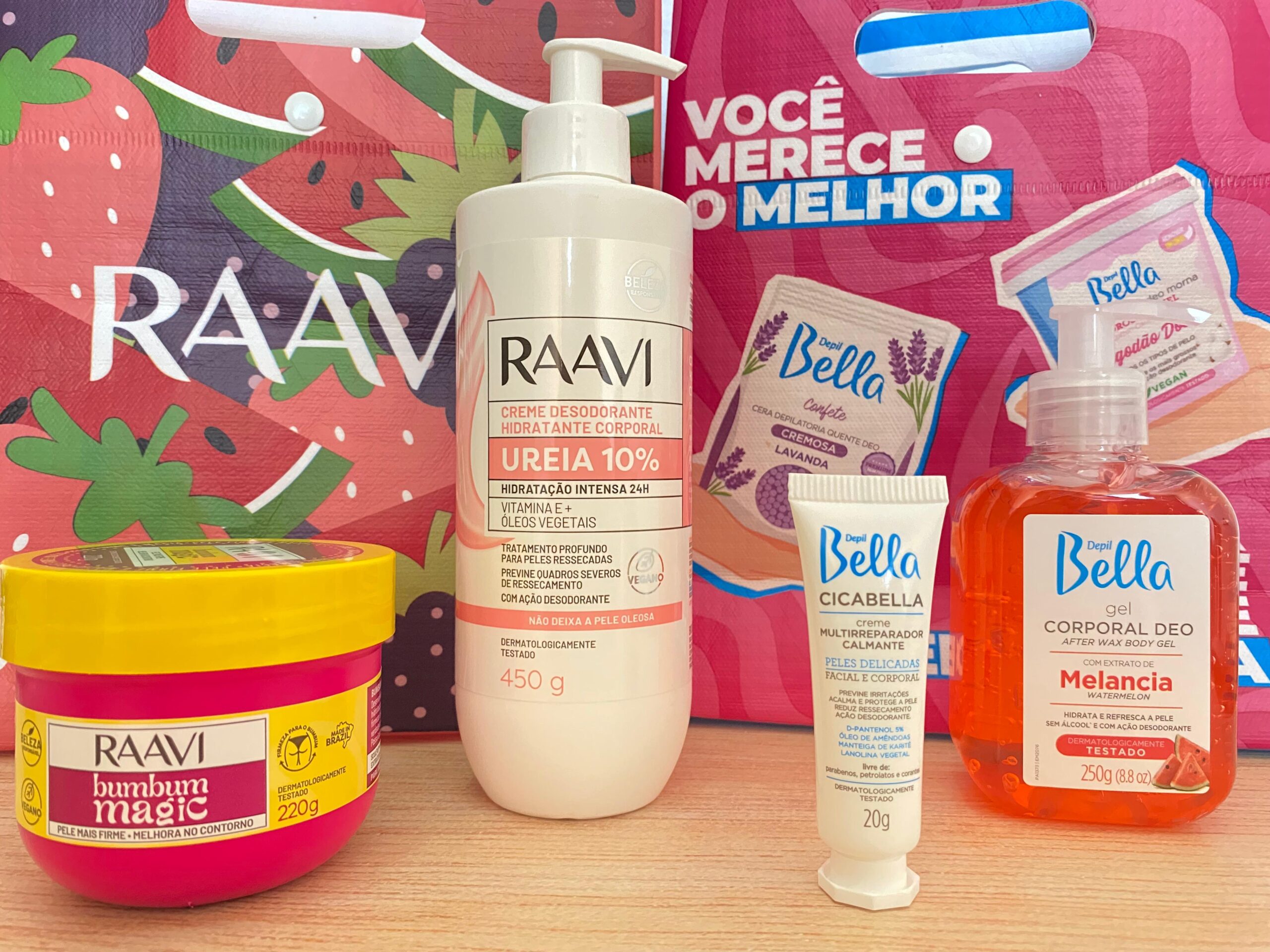 Produtos Depil Bella e Raavi exibidos juntos em frente às sacolas das marcas, com destaque para o hidratante corporal, máscara firmadora e gel pós-depilação apresentados na Beauty Fair 2025.