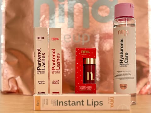 Produtos da Nina MakeUp expostos sobre mesa de madeira, incluindo Panthenol Lashes, Gloss Labial Instant Glow, Hyaluronic Care e lápis Instant Lips.