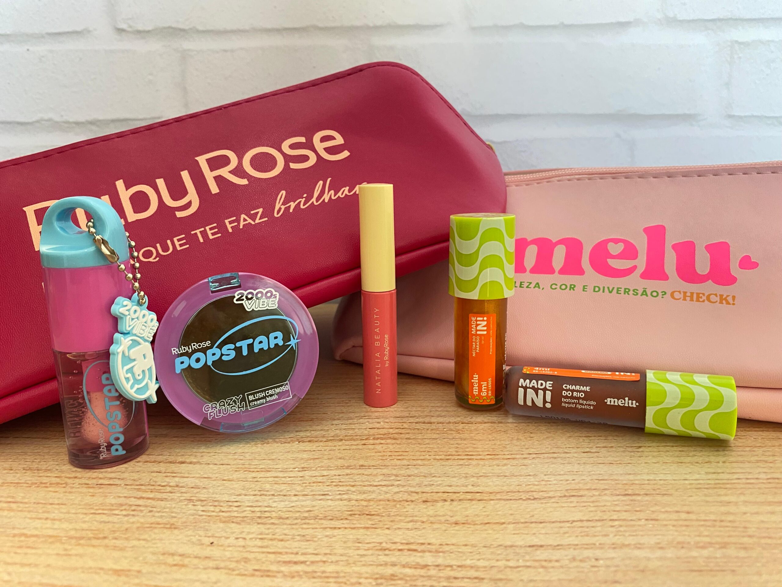 Produtos Ruby Rose e Melu sobre mesa de madeira com nécessaires das marcas, incluindo blush cremoso, gloss e batom líquido.