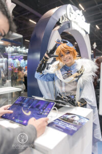 Visitante joga Genshin Impact em um tablet no estande da HoYoverse durante a BGS 2025, com cosplayer do personagem Childe interagindo com o público.