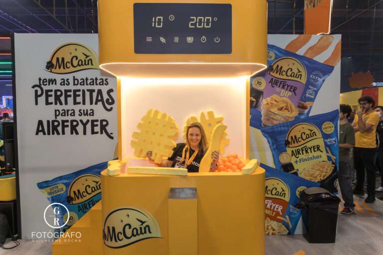 Visitante posa dentro de uma Air Fryer gigante no estande da McCain na BGS 2025, cercada por embalagens da linha AirFryer e batatas em formato divertido.