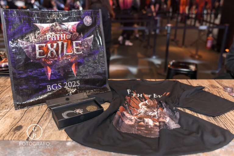 Brindes oficiais de Path of Exile 2 exibidos na BGS 2025, incluindo ecobag personalizada com o logo do jogo, camiseta temática preta e pin kit colecionável sobre uma mesa de madeira.