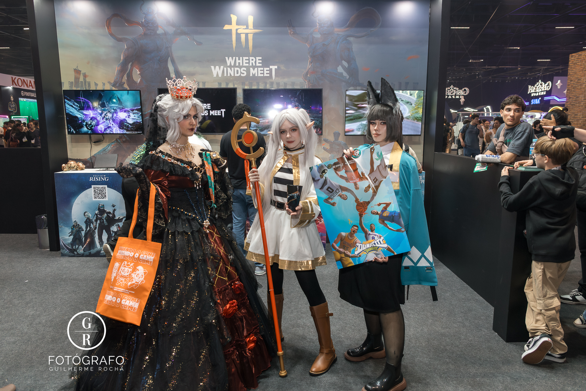 Cosplayers posam no estande da Smile One durante a BGS 2025, em frente à área dedicada aos jogos Where Winds Meet, Destiny Rising e Dunk City Dynasty, mostrando brindes e pôsteres temáticos oferecidos nas ativações da parceria com a NetEase.