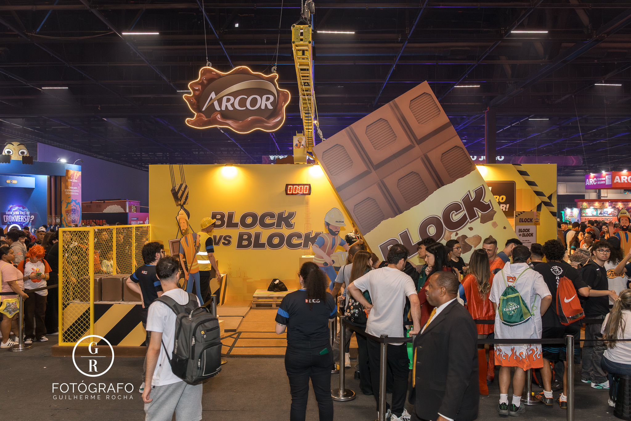 Estande da Arcor na Brasil Game Show com o tema “Block vs Block”, apresentando uma estrutura gigante em formato de barra de chocolate e área interativa cercada por visitantes, que participam de desafios e tiram fotos no espaço decorado com elementos da marca.