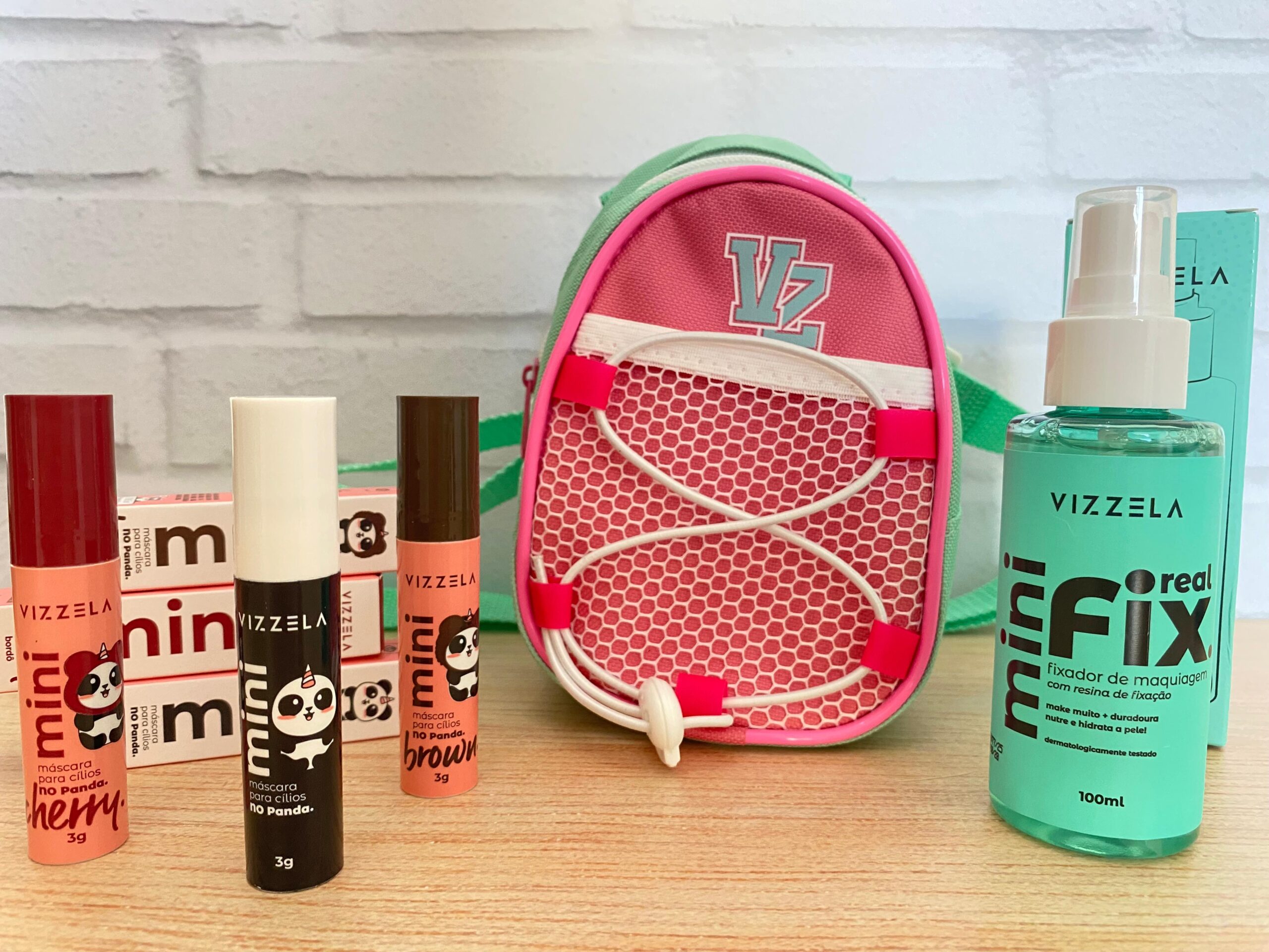 Kit Vizzela com máscaras de cílios Mini No Panda, fixador de maquiagem Mini Real Fix e mini mochila rosa e verde sobre fundo claro.