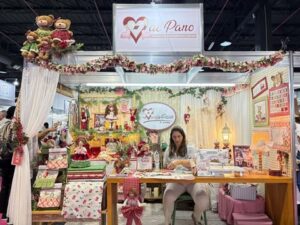 Estande decorado da marca Coraçãode Pano na feira Criativa, com artesã trabalhando e peças artesanais natalinas expostas.