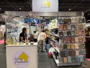 Visão lateral do estande da Casa da Madeira na Criativa Campinas, mostrando variedade de tecidos, tintas e materiais para artesanato.