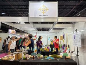 Estande Arte Brasil na Criativa, com peças artesanais coloridas expostas e visitantes conversando sobre os produtos.