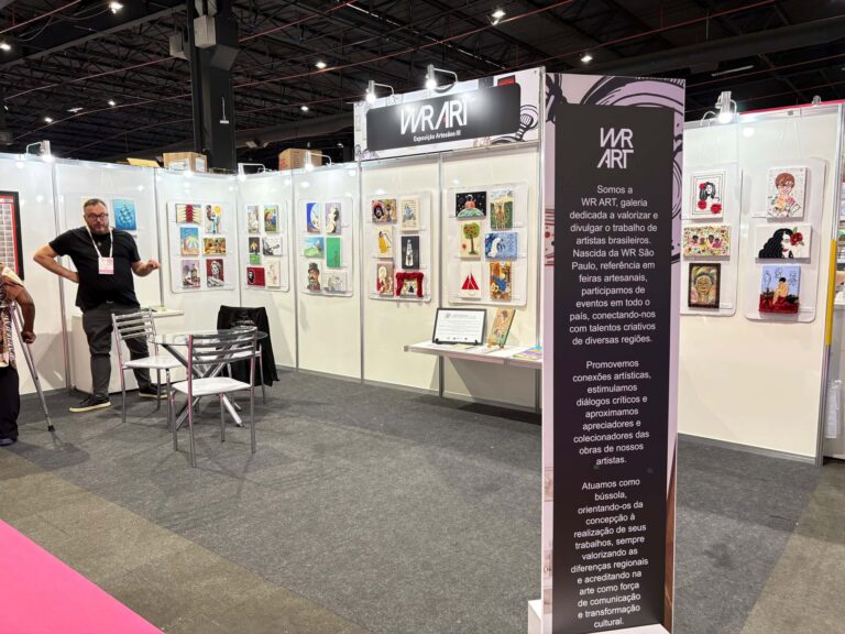Estande da WR Art na Criativa Campinas, com obras artesanais expostas nas paredes brancas, painel explicativo da galeria e visitantes observando o espaço.
