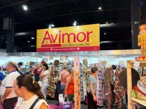 Estande da Avimor na Criativa, com grande variedade de tecidos estampados e visitantes circulando pelo corredor.