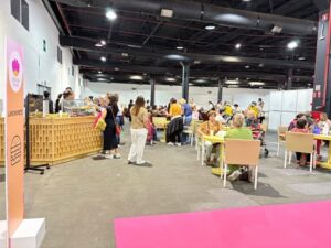 Área de alimentação da feira Criativa, com visitantes sentados às mesas, fila na lanchonete e espaço amplo iluminado.
