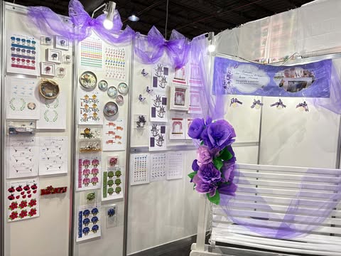 Painel de trabalhos artesanais na Criativa Campinas, com bordados, gráficos, recortes e flores decorativas em tons de roxo.