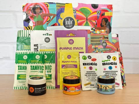 Amostras de produtos Lola dispostas sobre a mesa, incluindo sachês das linhas Tannic Acid, Purple Mask, Básicão e miniaturas de cremes de tratamento, todas em embalagens coloridas da marca.