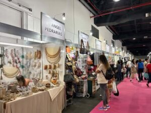 Corredor da Criativa com estandes de bijuterias e acessórios, incluindo o By Angel Pedrarias, exibindo peças artesanais para os visitantes.