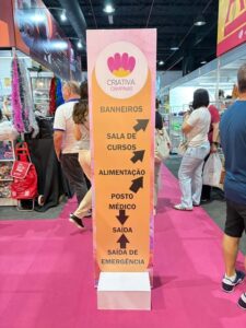 Totem informativo da Criativa exibindo setas para banheiros, sala de cursos, alimentação, posto médico, saída e emergência.
