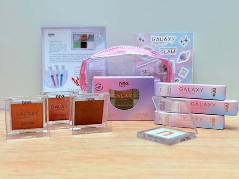 Coleção Galaxy Nina MakeUp completa com glosses, paleta de sombras, blushes e iluminador organizados sobre superfície de madeira.