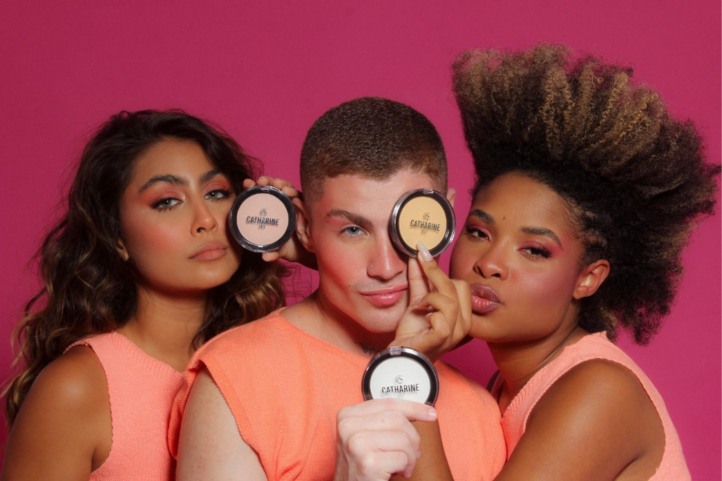 Três modelos exibem o pó translúcido da Catharine Hill em frente a um fundo rosa, destacando diversidade e acabamento uniforme da maquiagem.