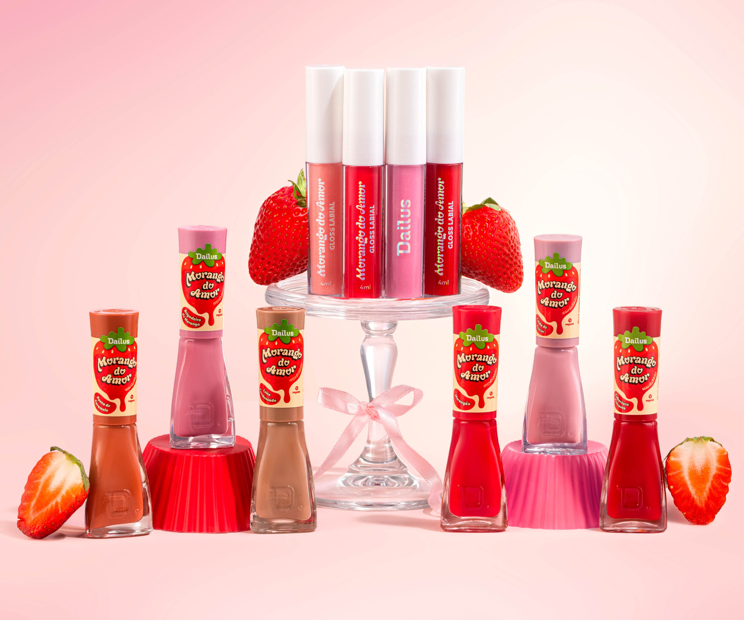 Coleção Morango do Amor da Dailus com glosses labiais e esmaltes expostos em suporte de vidro, acompanhados de morangos frescos sobre fundo rosa.