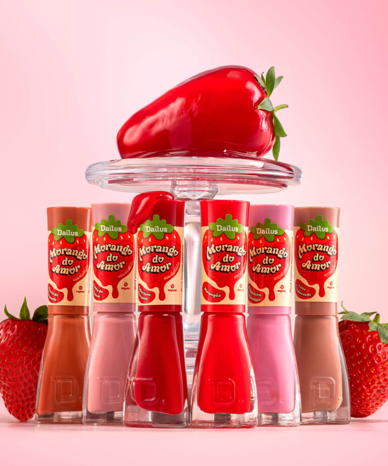 Esmaltes Morango do Amor da Dailus em seis cores, exibidos sobre suporte transparente com um grande morango decorativo, em fundo rosa vibrante.