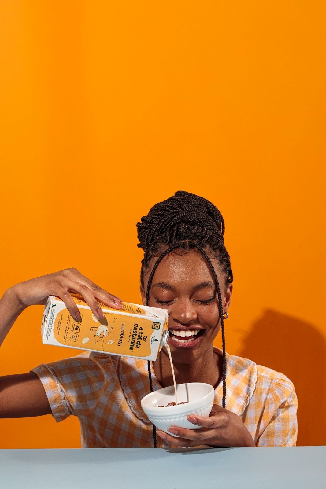 Jovem sorrindo enquanto despeja bebida vegetal A Tal Da Castanha em uma tigela de cereal, em frente a um fundo laranja vibrante.