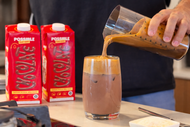 Bebida gelada de chocolate vegetal é servida em copo com gelo, com embalagens de Possible ao fundo, destacando preparo cremoso e refrescante.
